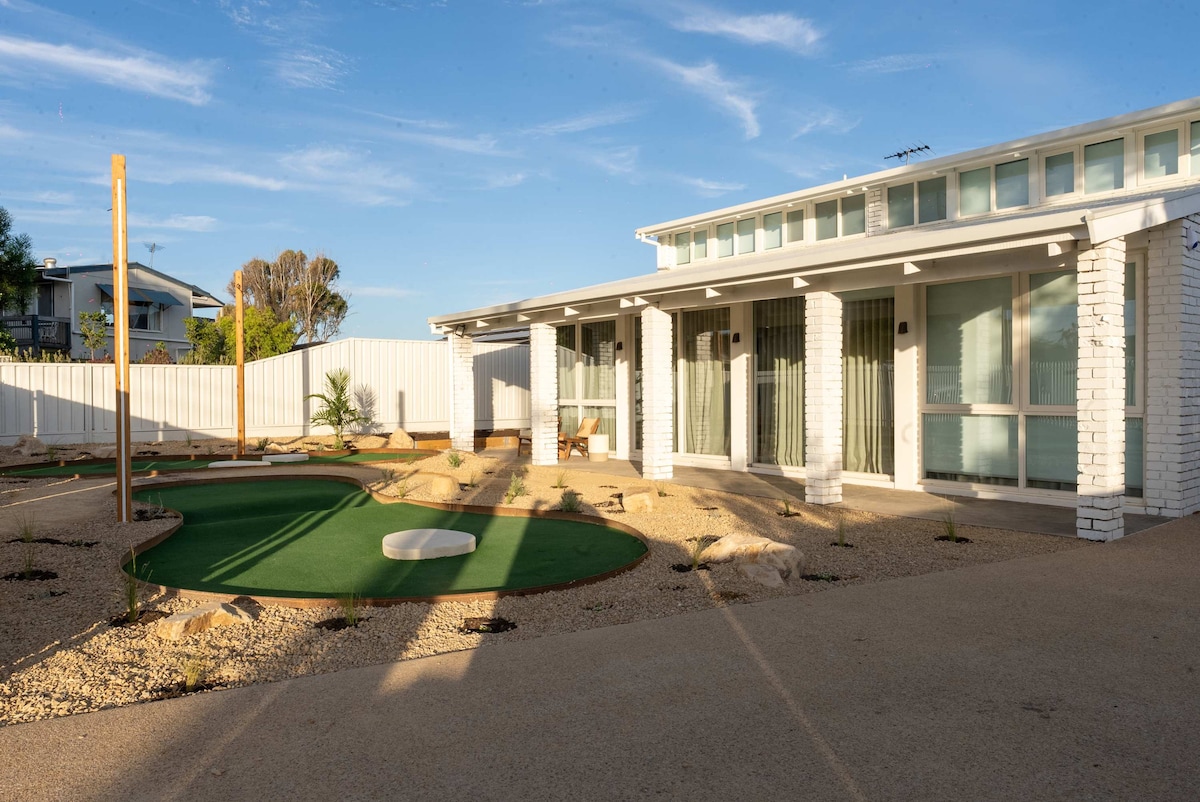 The exterior of the home is showcased, featuring a landscaped yard with a fun putt-putt area. The light-colored building has large windows reflecting the sky, and the surrounding space includes minimalistic landscaping with gravel and a few plants.