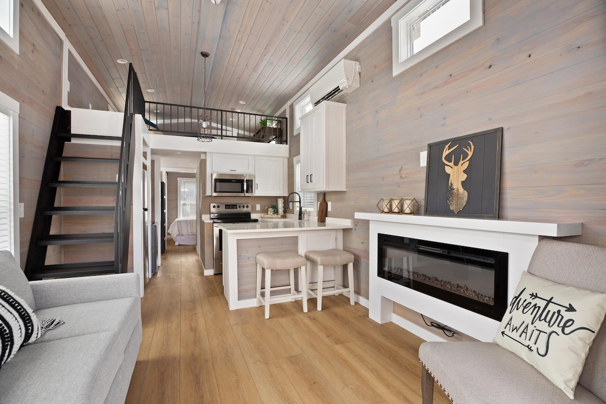 An open-concept living area showcases light wood paneling and a spacious kitchen with modern appliances. Two stools are positioned at the kitchen island, complemented by a cozy seating area featuring a pull-out couch. Natural light filters through windows, enhancing the inviting ambiance.