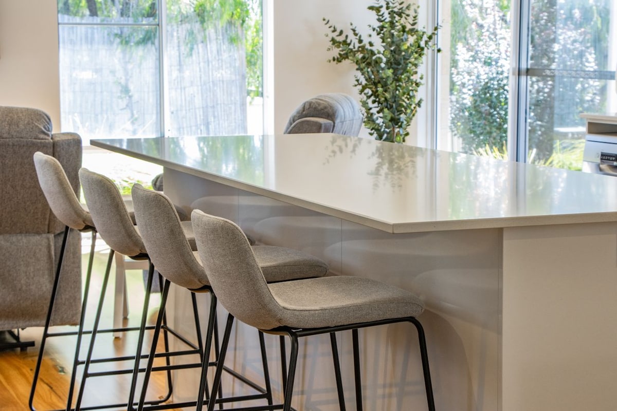 A modern kitchen island is highlighted, featuring four soft grey bar stools with black metal frames. Natural light floods the space through large windows, accentuating the clean lines of the sleek countertop and the greenery outside.