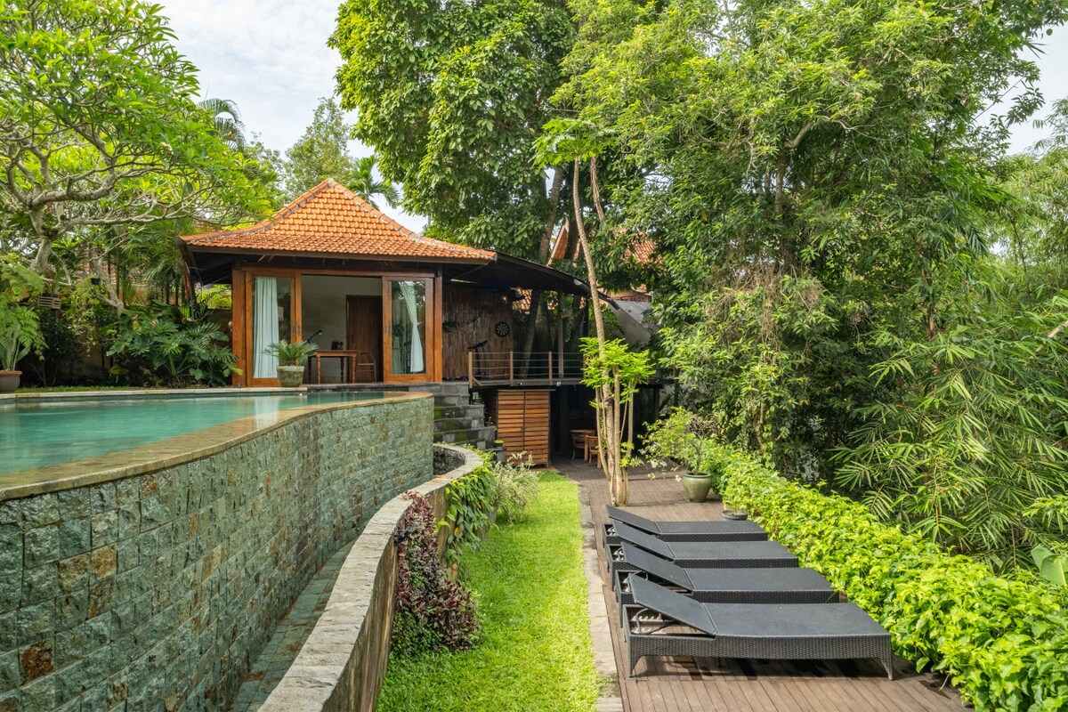 A serene infinity pool is bordered by lush greenery, with modern lounge chairs positioned along the poolside. Nearby, a traditional roofed structure emerges among the trees, complementing the tranquil atmosphere of the garden path leading to it.