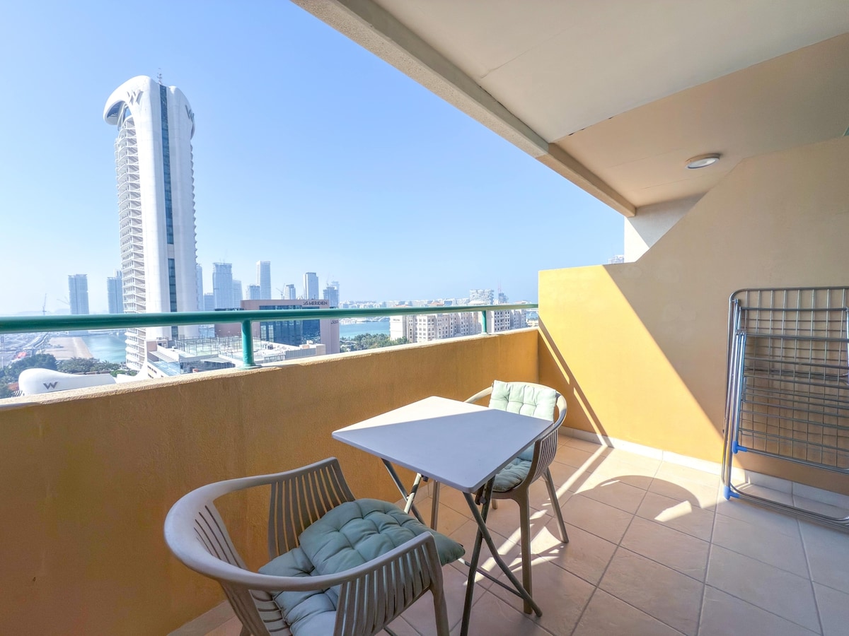 A private balcony features a small table and two comfortable chairs, surrounded by a clear view of the city skyline and the iconic tower structures. Natural light brightens the space, highlighting the balcony's neutral-colored walls and tiled flooring.