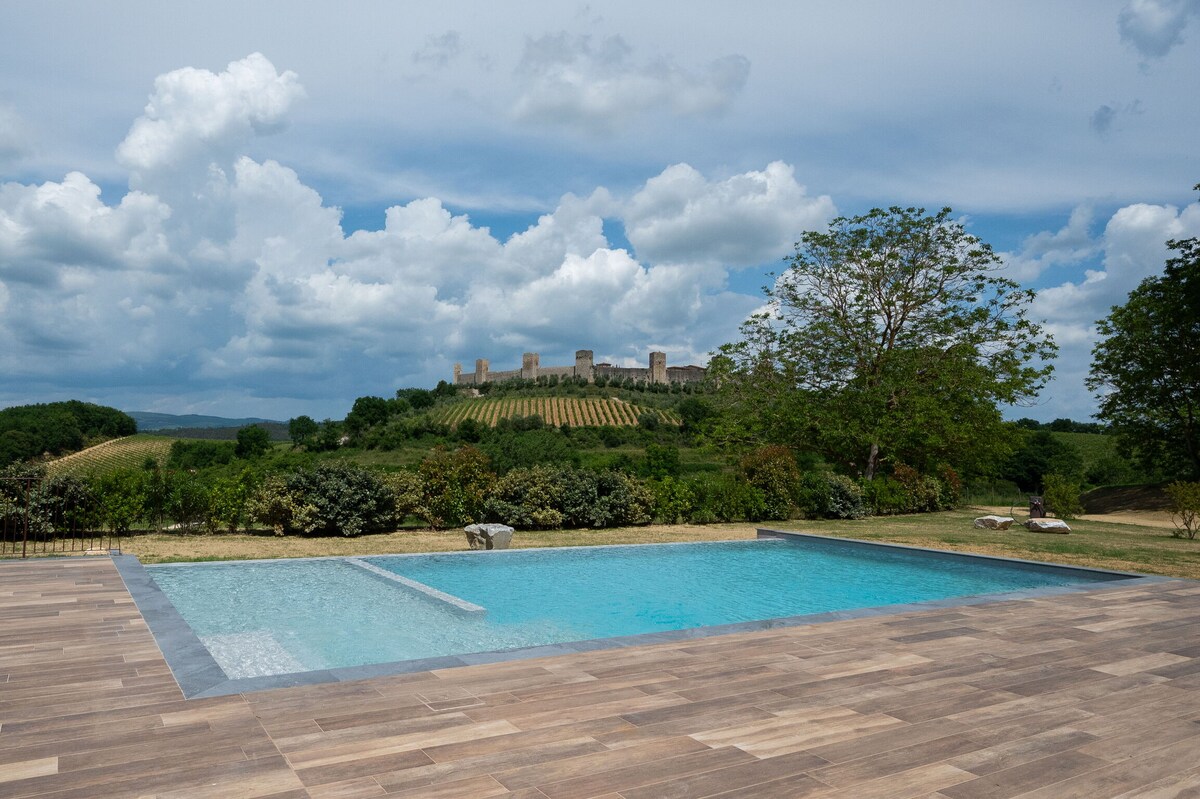 A clear view of a shared swimming pool is presented, with light blue water reflecting the sky above. The surrounding area is framed by lush greenery and vineyards. In the background, the medieval towers of Monteriggioni castle rise against a cloudy sky.