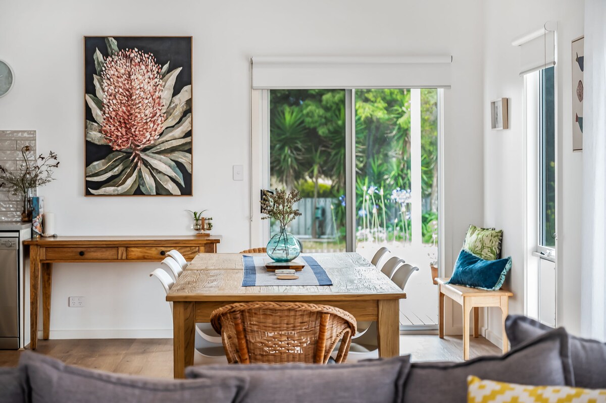 A dining area features a wooden table surrounded by white chairs, complemented by a rattan seat. A large artwork depicting a native flower adorns the wall. Natural light floods the space, highlighting views of a lush garden visible through expansive sliding glass doors.