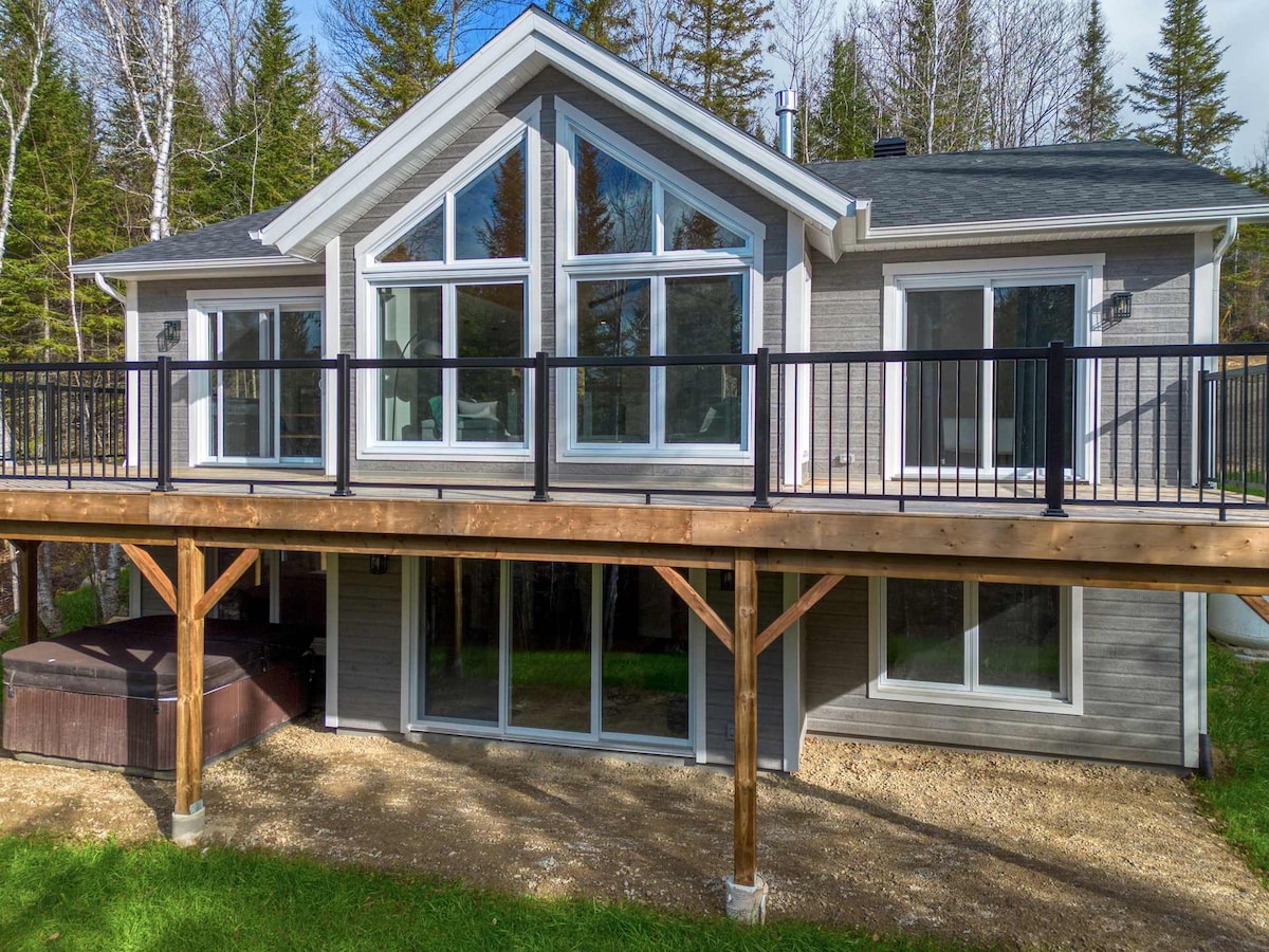 The exterior of the chalet features a large deck supported by wooden posts, providing access to the entrance. Large windows are visible, allowing natural light into the interior, while the hot tub is situated on the deck beneath a protected overhang.