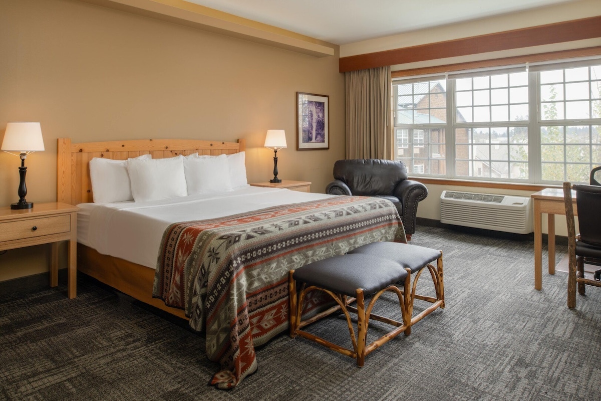 A spacious guestroom showcases a king bed adorned with decorative bedding, complemented by bedside lamps on wooden tables. A comfortable chair and work desk are visible, while natural light streams through a large window, offering views of the surroundings.