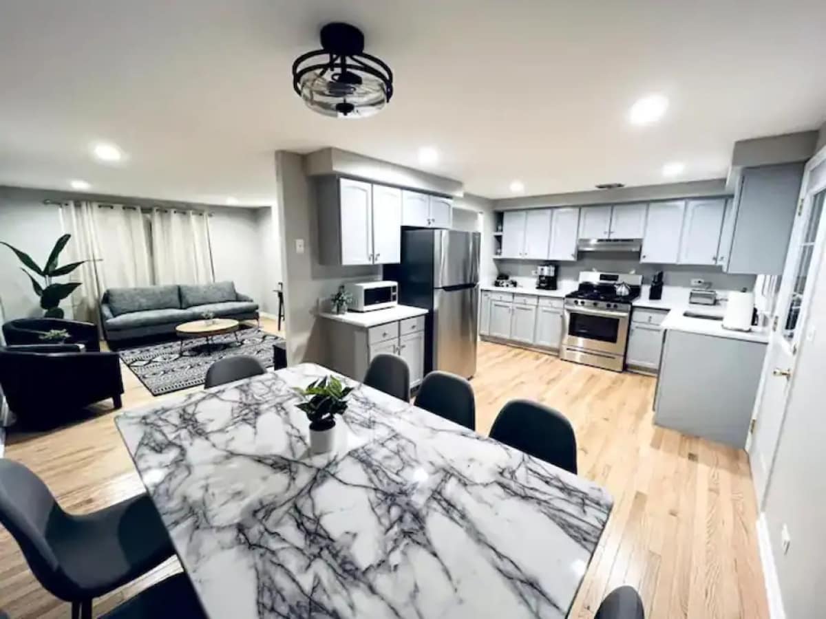 A modern kitchen area features light grey cabinetry and stainless steel appliances, including a refrigerator and stove. A marble dining table, surrounded by grey chairs, is positioned in front of a cozy seating area with a sofa and a textured rug. Natural light fills the space.