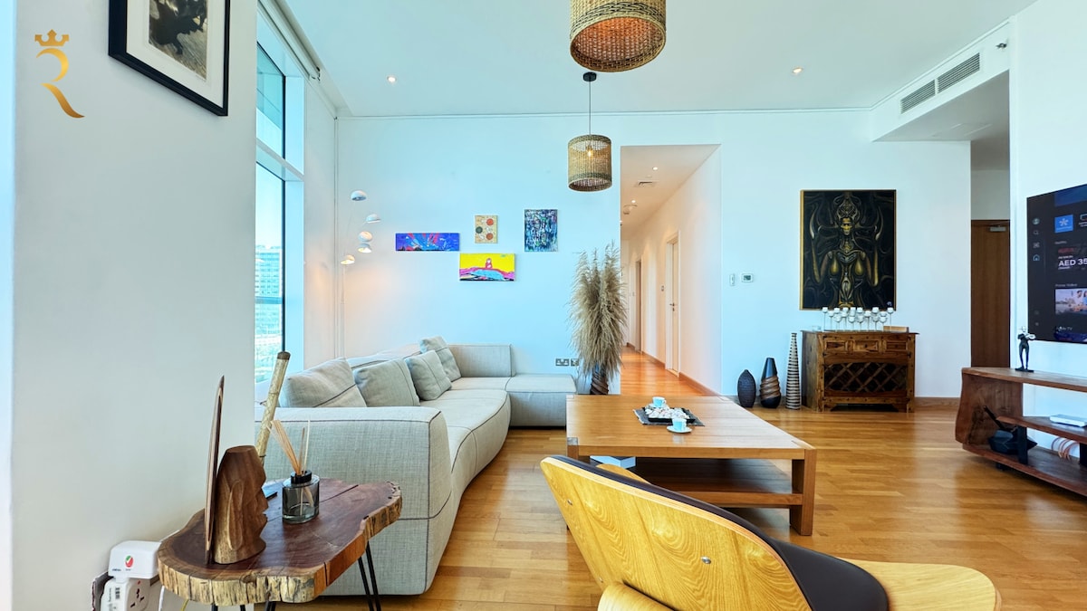A spacious living area features large windows providing ample natural light and stunning ocean views. A stylish L-shaped sofa invites relaxation, complemented by an elegant wooden coffee table. Art pieces adorn the walls, while modern decor and lighting enhance the inviting ambiance.