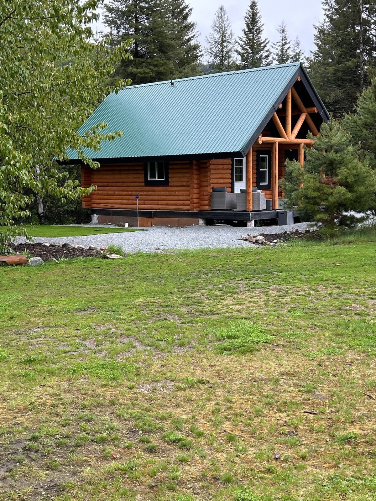 A charming log cabin is set within a lush green landscape, featuring a metal roof and log exterior. The entrance is framed by wooden beams, and a small porch with seating is visible. Surrounding trees add to the tranquil setting.