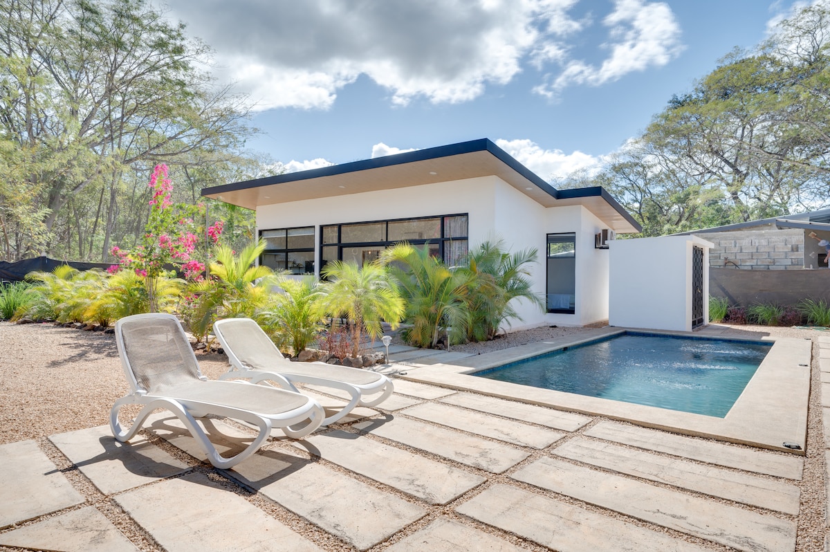 The exterior of the house is featured, showcasing a modern design with large windows and a welcoming pool. Two lounge chairs are positioned near the private in-ground pool, surrounded by lush landscaping and tropical plants. The setting under a partly cloudy sky offers a serene atmosphere.