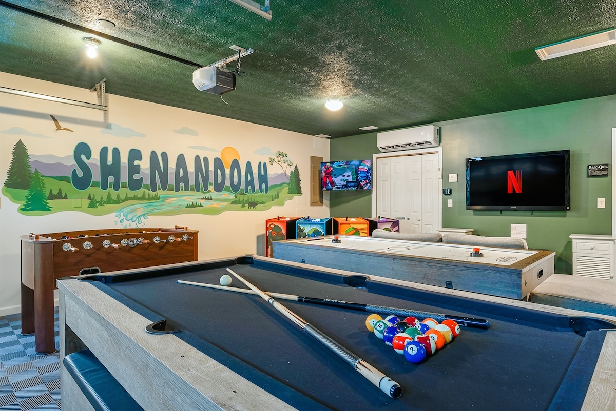 The game room is furnished with a multi-functional pool and ping-pong table, alongside an arcade console displaying various game options. A large mural of Shenandoah decorates the wall, and a television is mounted for entertainment viewing.