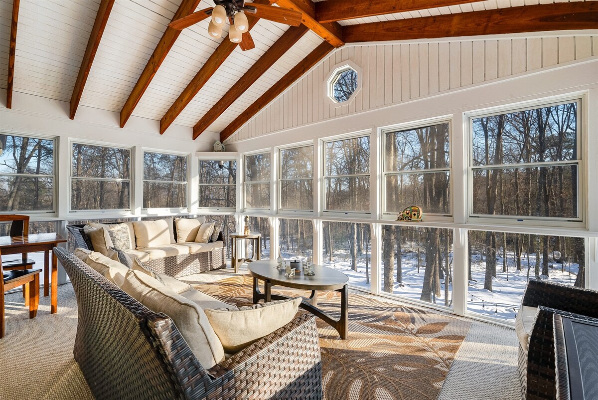 A sunlit sunroom features large windows framed with wood, offering views of a winter landscape. Comfortable seating arrangements are present, with a light-colored sofa and wicker chairs surrounding a central coffee table. The ceiling has exposed wooden beams, adding a rustic touch.