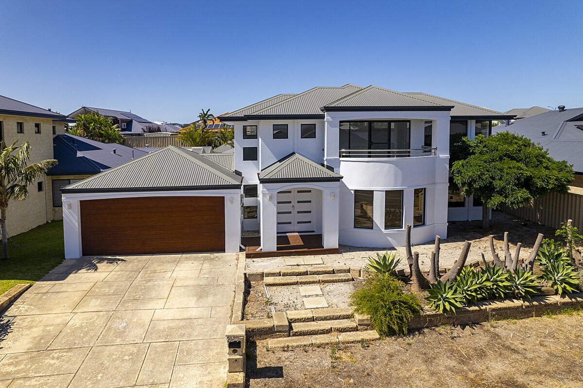 An expansive two-story home is showcased from an aerial view, featuring a modern façade with varying window sizes. A well-maintained front yard includes a pathway and manicured landscaping. The attached garage offers space for vehicle parking, while clear blue skies provide a bright backdrop.