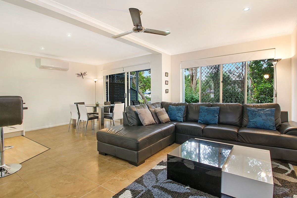 A spacious living area is furnished with a large black sectional sofa adorned with decorative cushions. A glass coffee table sits on a patterned rug, and a dining space with a round table and chairs is visible in the background. Large windows allow natural light to fill the room.
