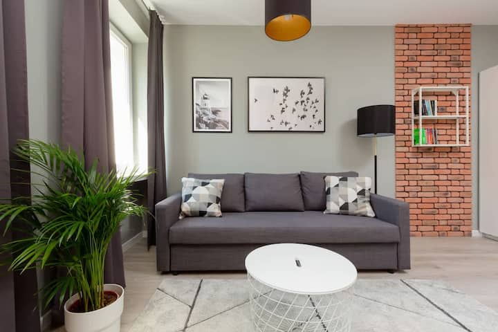 Un salon moderne avec un canapé gris foncé, une table basse ronde blanche et un mur d'accent briques, complété par des œuvres d'art décoratives et une plante en pot.