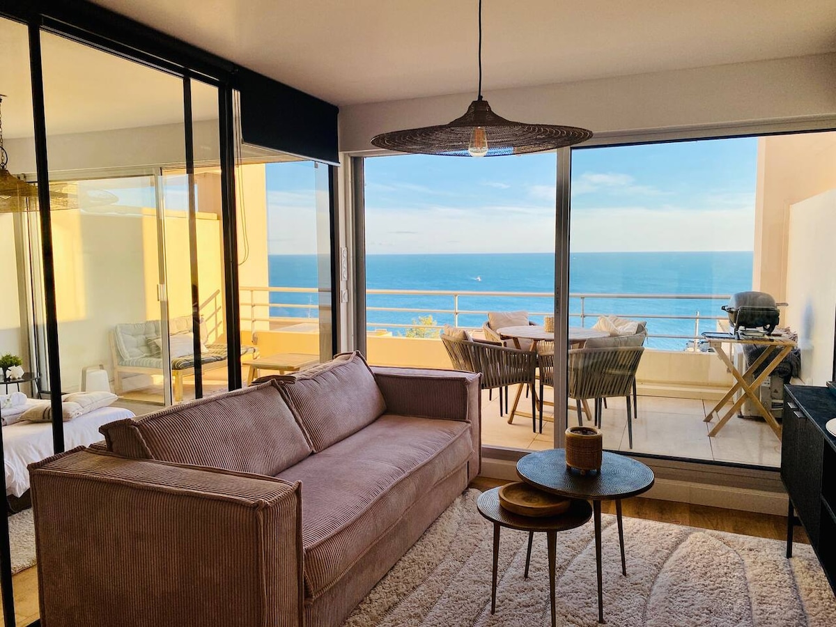 An inviting living space is visible, featuring a plush sofa facing large glass doors that open to a terrace with ocean views. A dining area with a table and outdoor seating is also seen, along with a view of the sea and a barbecue grill.