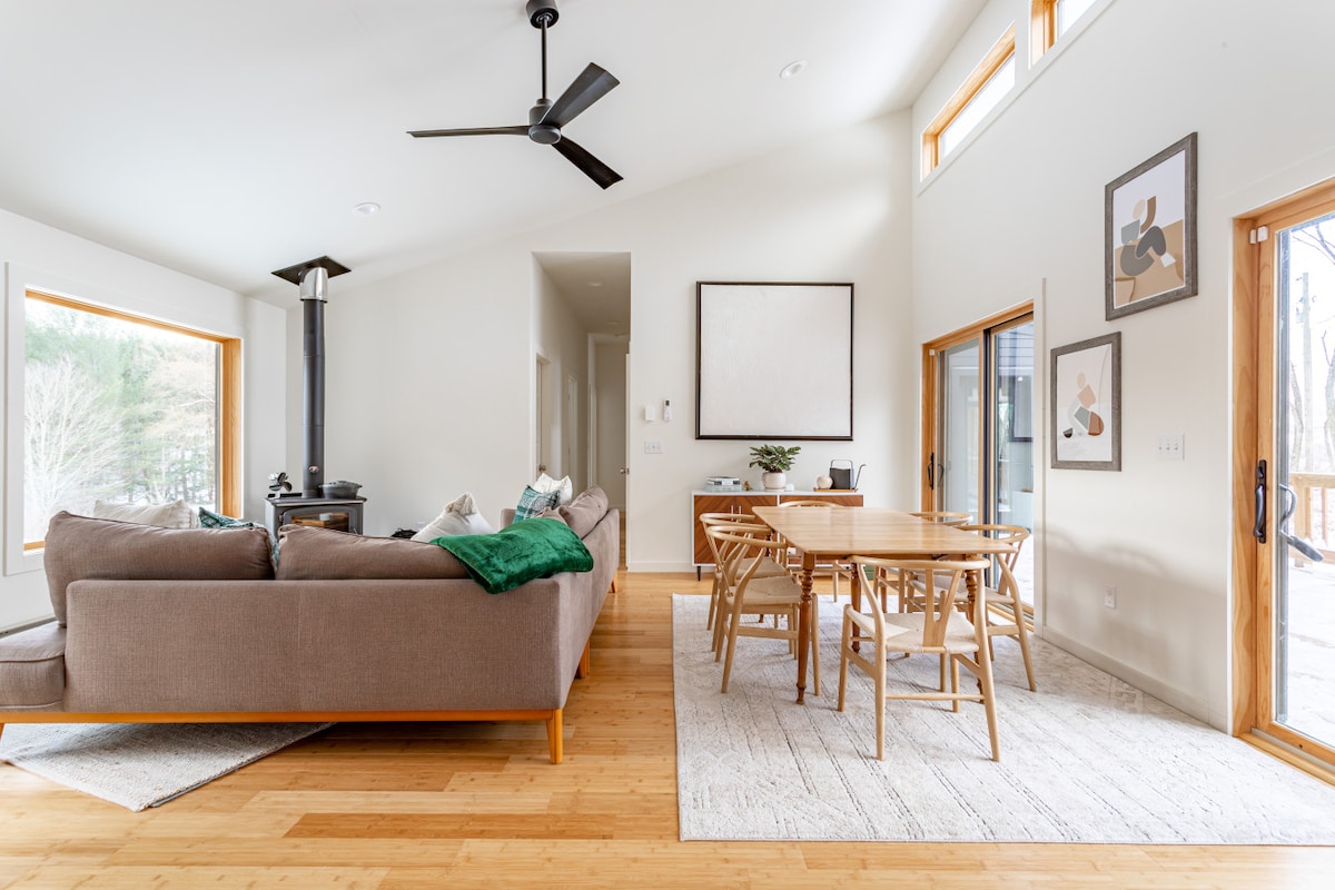 A spacious living area features a large sectional sofa adorned with a green throw blanket, complemented by a wooden dining table with seating for six. Natural light floods in through multiple large windows, and a wood-stove stands as a cozy centerpiece against the white walls.
