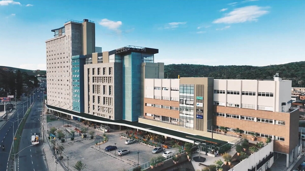 The image captures a modern multi-story building featuring a mix of architectural styles. Large windows are visible on the front, allowing natural light. A landscaped area with parking spaces is noted in the foreground, alongside well-maintained pathways. The surrounding hills provide a scenic backdrop.