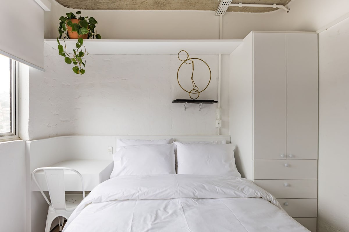 A bright sleeping area features a spacious bed with fresh white linens. A small desk is positioned beside the bed, accompanied by a white chair. A decorative plant rests on a shelf above, adding a touch of greenery to the minimalist design.
