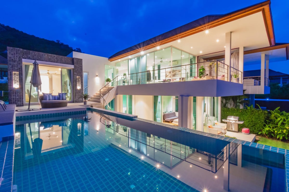 A modern two-story villa is illuminated at dusk, featuring large glass sliding doors that create a seamless connection between the living area and the outdoor space. The inviting swimming pool reflects the villa's structure, enhancing the serene atmosphere of the secluded setting.