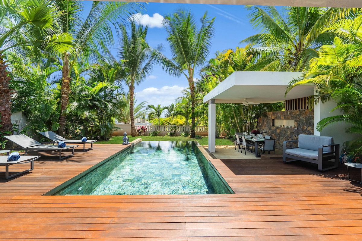 An expansive outdoor space features an infinity pool surrounded by lush tropical plants and palm trees. Comfortable sun loungers are positioned along the wooden deck, while a covered dining area sits nearby, furnished with a table and chairs for outdoor meals.