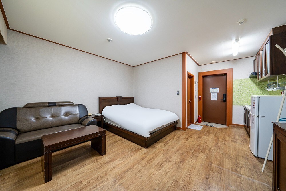 The room features a spacious bed with a simple headboard, accompanied by a comfortable sofa and a wooden coffee table. A kitchenette is visible, along with a refrigerator and a bright green backsplash. The entrance door and a large window contribute to the overall functionality of the space.