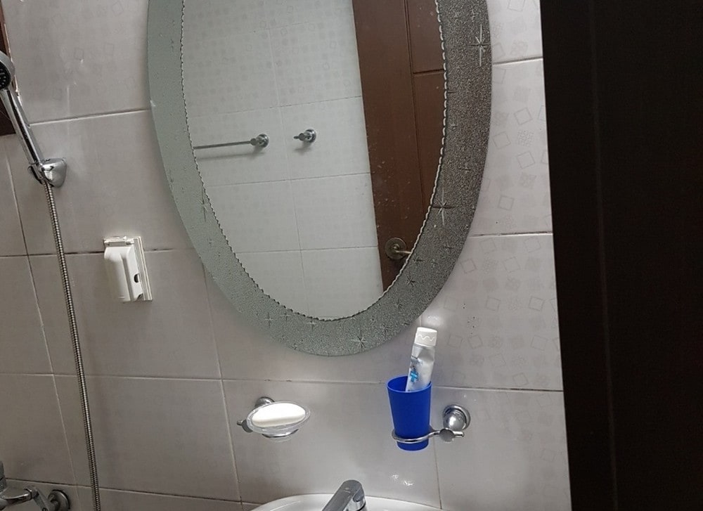 A bathroom is depicted, featuring a round mirror above a sink. The sink is accompanied by a wall-mounted soap dispenser and a blue cup. Clean tiles are visible on the walls, with shower fixtures mounted nearby, highlighting a functional and tidy space.