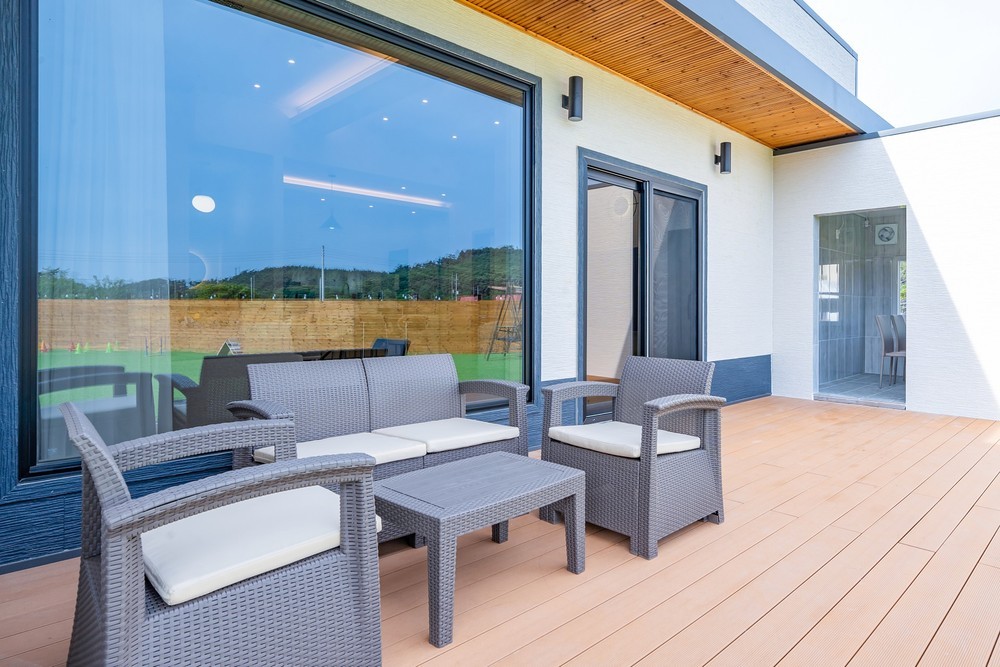 An outdoor patio area is presented with wicker chairs and a low table, furnished with plush white cushions. Large windows offer a view of the surrounding greenery, while wooden decking enhances the serene environment.
