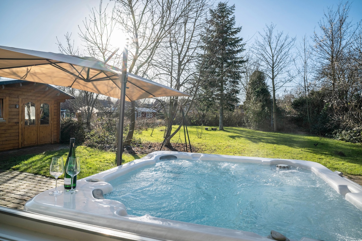 A private whirlpool is set against a backdrop of a well-maintained garden, with a soft canopy providing shade. Clear skies and sunlight enhance the serene atmosphere, with trees and greenery framing the relaxing outdoor space.
