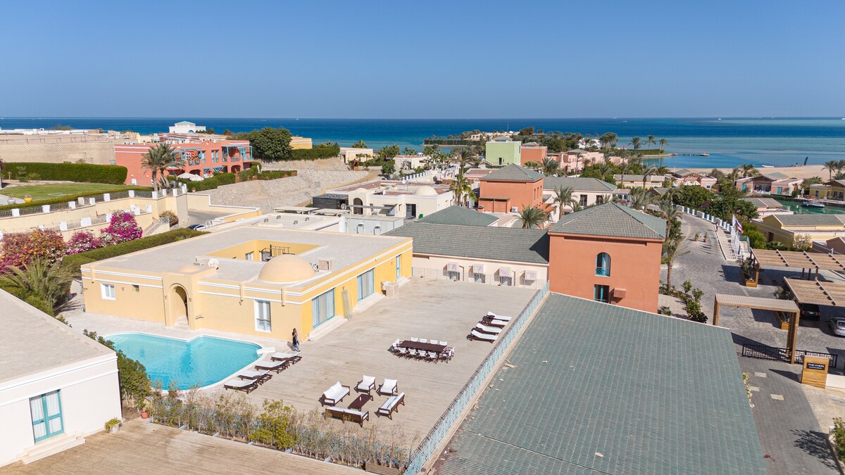 An aerial view captures a sprawling villa complex, featuring a clear blue pool surrounded by sun loungers. The distant sea glimmers under sunlight, with colorful buildings and lush greenery framing the landscape, highlighting the inviting outdoor space of this luxurious getaway.