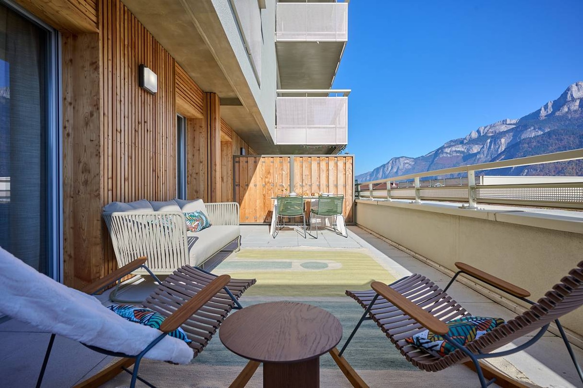 A spacious terrace features modern outdoor furniture, including two lounge chairs and a small wooden table. The setting offers a panoramic view of the surrounding mountains, with clear skies enhancing the serene atmosphere. A dining area is also present, accommodating up to four guests.