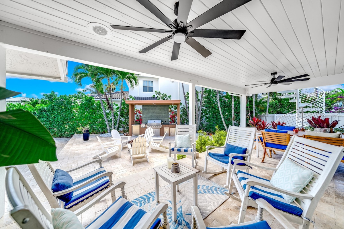 A covered outdoor seating area features several white lounge chairs with blue cushions, providing comfort for relaxation. The space includes ceiling fans for airflow, with lush greenery surrounding an outdoor kitchen in the background, enhancing the inviting atmosphere.