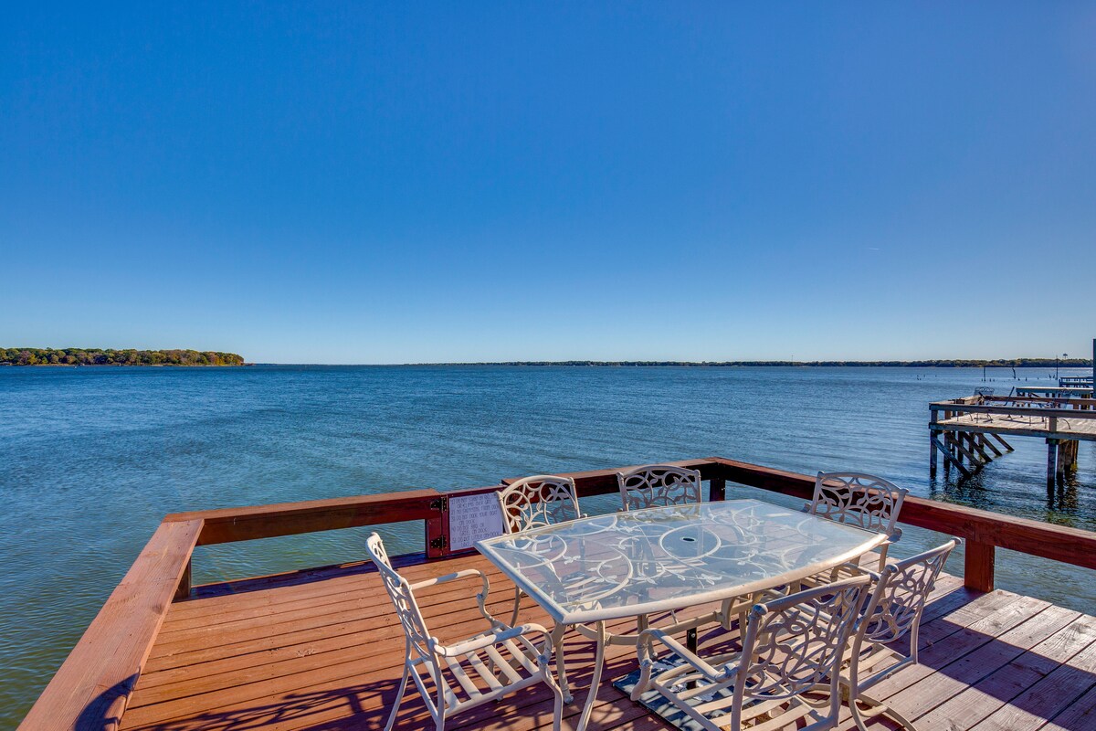 A lakeside dock features a large glass-top table surrounded by six decorative metal chairs, providing a space for dining while enjoying waterfront views. The calm water stretches into the distance under a clear blue sky, creating a serene atmosphere.