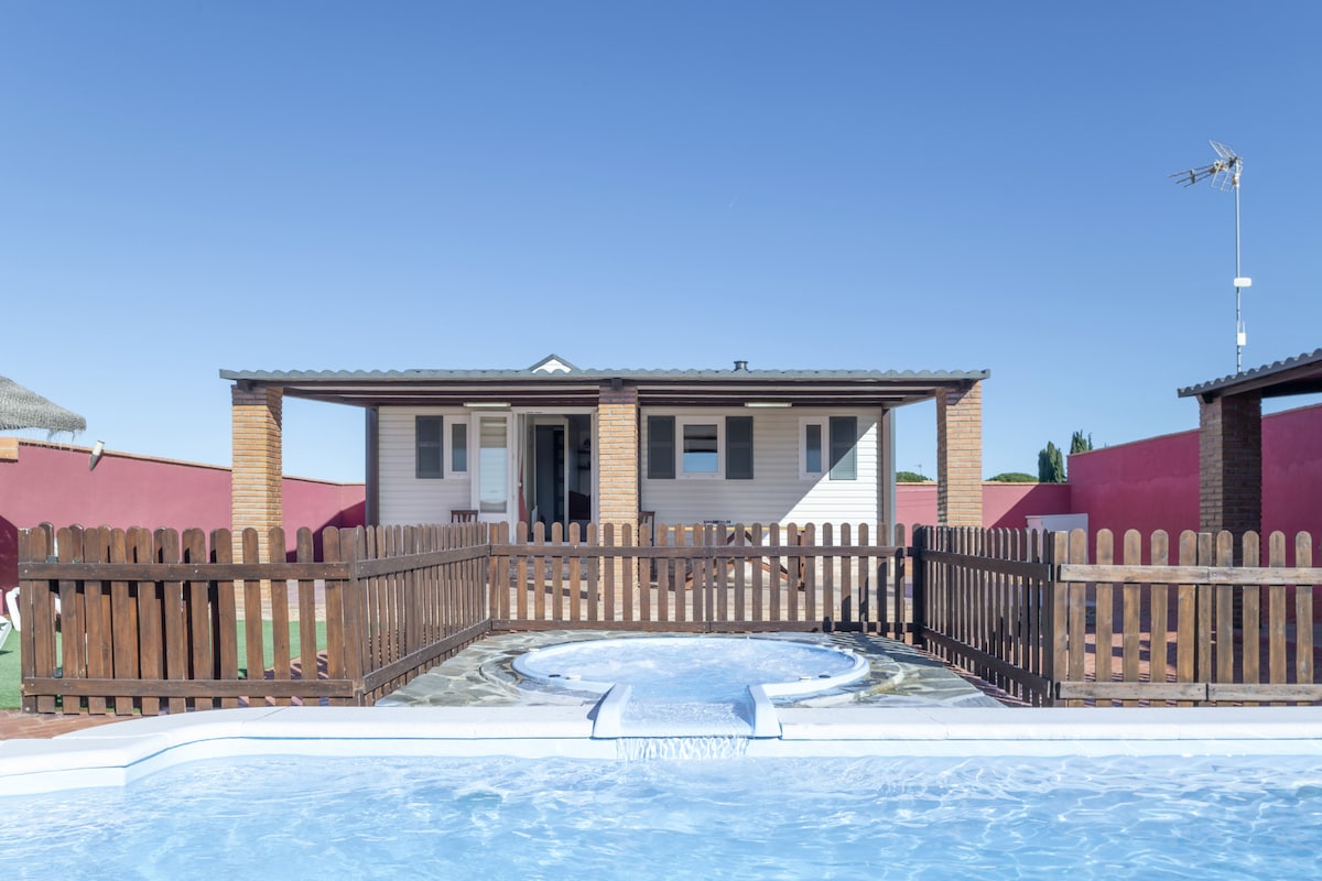 The outdoor area showcases a private pool accompanied by a whirlpool, both surrounded by a wooden fence. A shaded seating area is visible nearby, with the bungalow in the background and clear blue skies above.