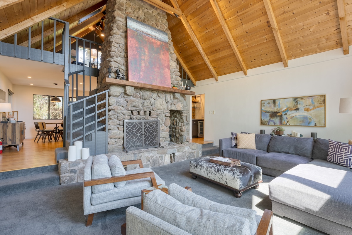 A spacious living area features a prominent stone fireplace, framed by vaulted pine ceilings and large windows offering natural light. Comfortable gray seating arrangements include a large sectional and accent chairs, while decorative art pieces and soft textiles contribute to the inviting ambiance.