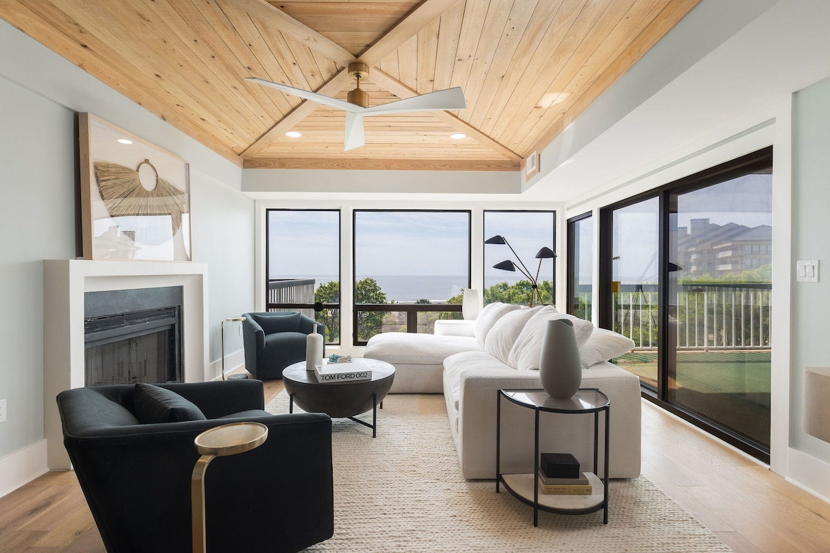 The living area showcases wide plank engineered hardwood floors and a stunning cypress ceiling, emphasizing natural materials. Large windows invite natural light and offer serene ocean views. Comfortable seating includes a sofa and two chairs, complemented by a circular coffee table and decorative accents.