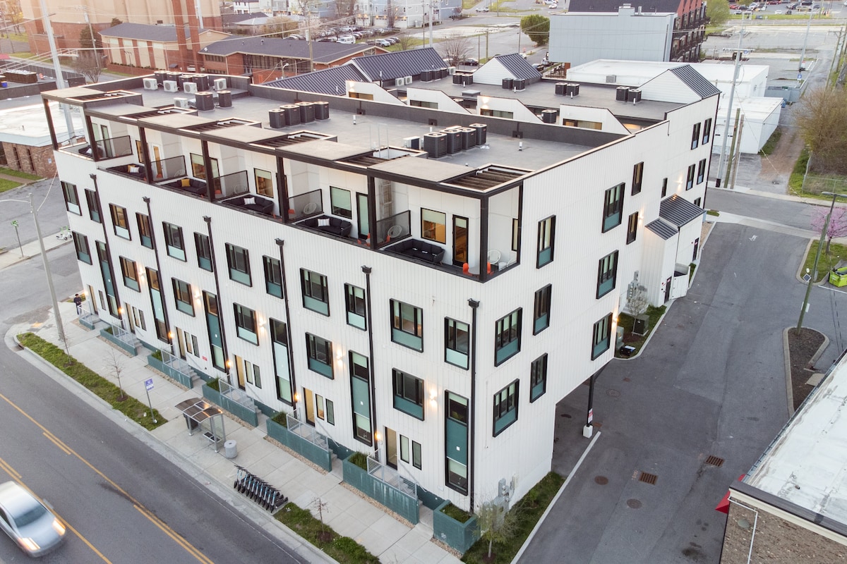 The exterior image showcases a contemporary multi-story building with a white façade and large windows. Balconies adorn the upper levels, providing outdoor space. The surrounding area features landscaped greenery and nearby streets, indicating a vibrant urban neighborhood.