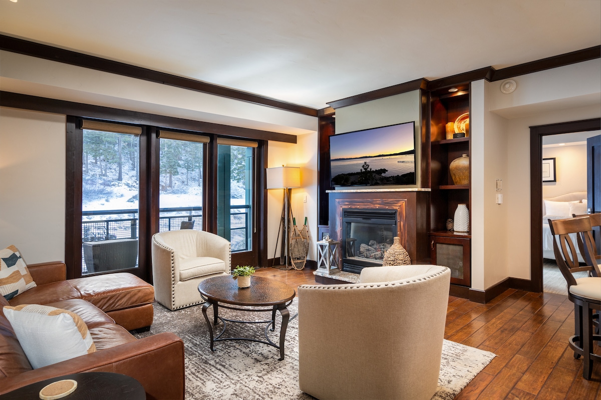 The living room is designed with an open floor plan, featuring a cozy gas fireplace surrounded by dark wood accents. Soft seating includes a leather sofa and two accent chairs. Natural light fills the space through multiple large windows, offering views of the outdoors.