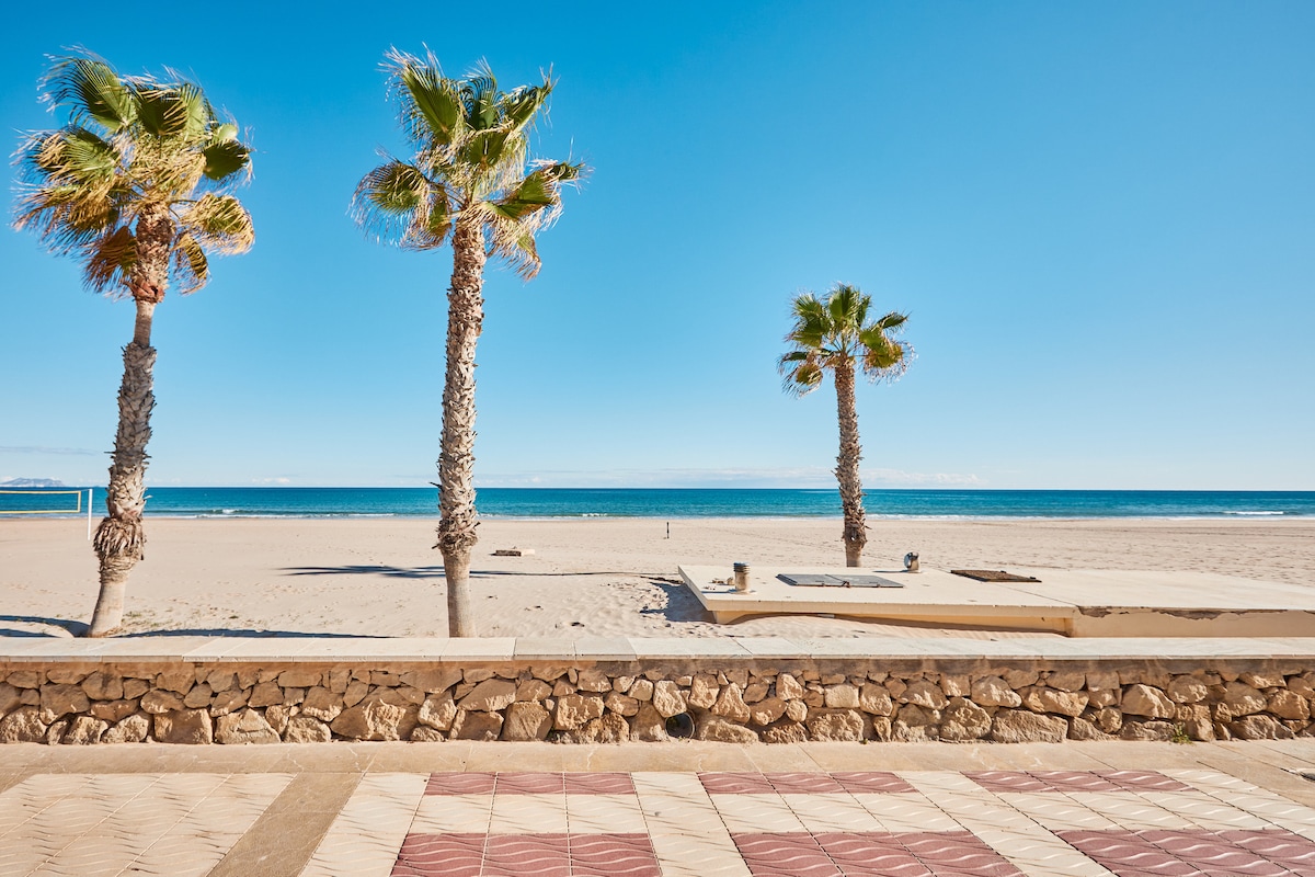 A coastal view is captured, highlighting the soft sandy beach and clear blue waters. Three palm trees are positioned in the foreground, swaying gently against a bright sky. A stone wall and patterned tile floor are visible, adding context to the tranquil seaside setting.