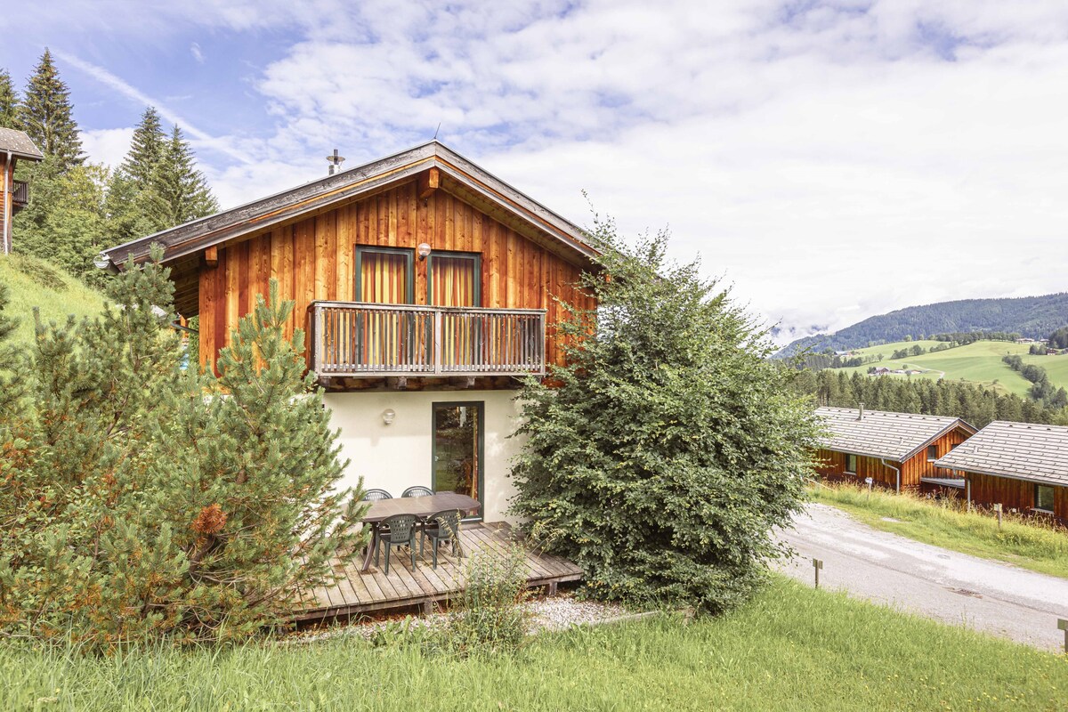 A wooden chalet is positioned amidst lush greenery, featuring a balcony that overlooks scenic hills. A small outdoor seating area with a table and chairs is visible on the furnished deck, set against a backdrop of trees and mountains.
