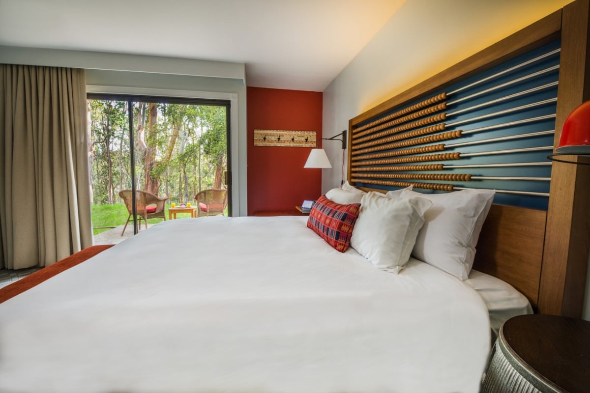 A cozy room features a spacious king bed adorned with soft linens and pillows. A large sliding glass door opens to a patio, surrounded by trees. Soft lighting highlights the wood-paneled headboard and a small seating area with two chairs.