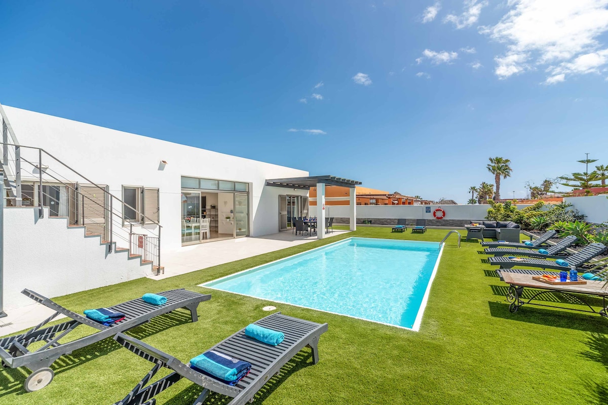 A clear view of the private pool surrounded by artificial grass and sun loungers. The bright blue water reflects the sky, while a shaded area near the villa adds to the outdoor space. A few palms can be seen in the background against a bright blue sky.