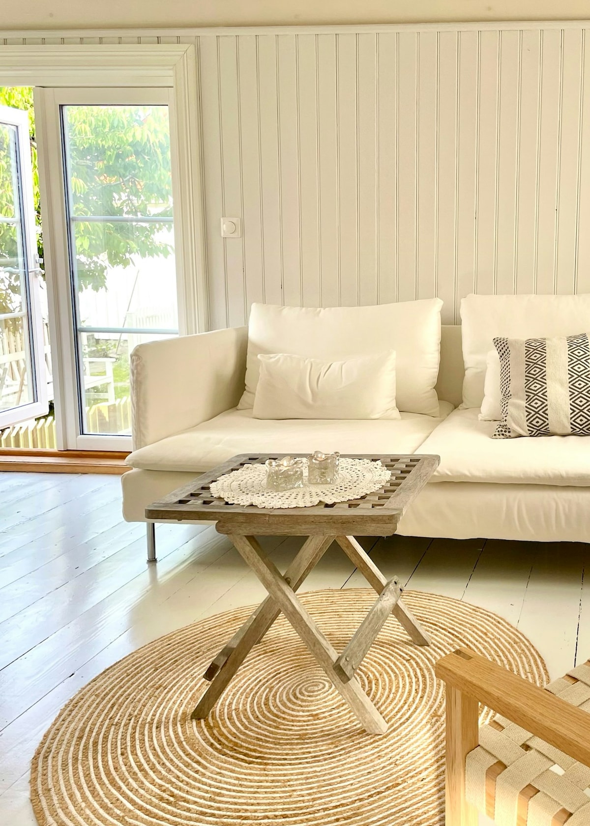 A light and airy living space features a white sofa adorned with patterned cushions. A rustic wooden coffee table rests on a round jute rug, adding texture to the room. Large glass doors open to the patio, allowing natural light to fill the space.