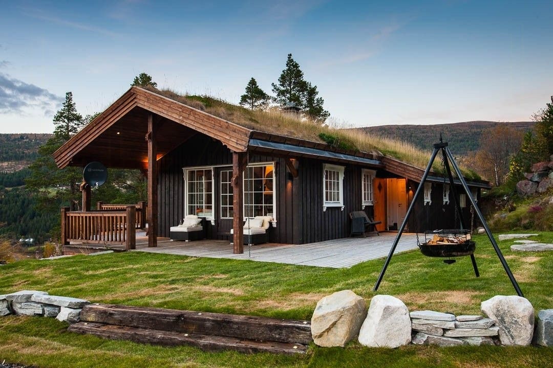 A cozy cabin with a sloped roof is surrounded by greenery, showcasing a spacious patio area. Outdoor furniture is placed on the lawn, alongside a fire pit, framed by natural stone. Large windows allow for plenty of light, enhancing the inviting exterior.