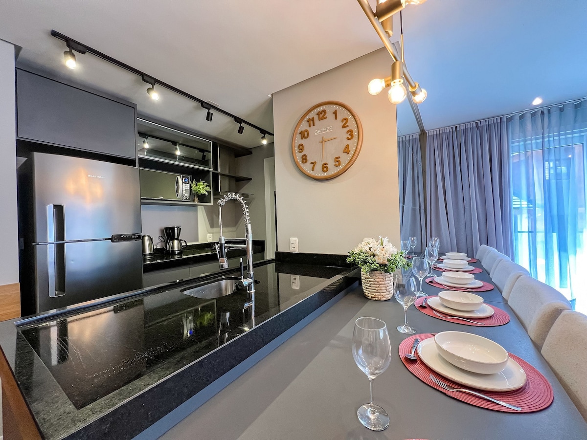 A modern kitchen area is presented with sleek black countertops and minimalist cabinetry. A round clock adorns the wall, complementing the stylish dining setup. A long table is set for six, featuring elegant plates and glassware, with natural light filtering through sheer curtains.