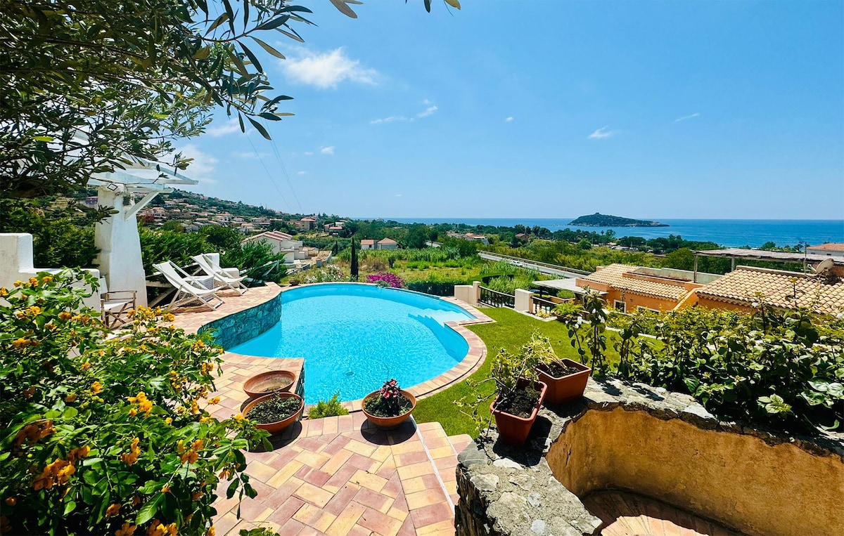A view of a private pool surrounded by lush greenery and terracotta tiles. Sun loungers are arranged beside the pool, which reflects the clear blue sky. The landscape features vibrant flowers and rolling hills, with the glittering sea visible in the distance.
