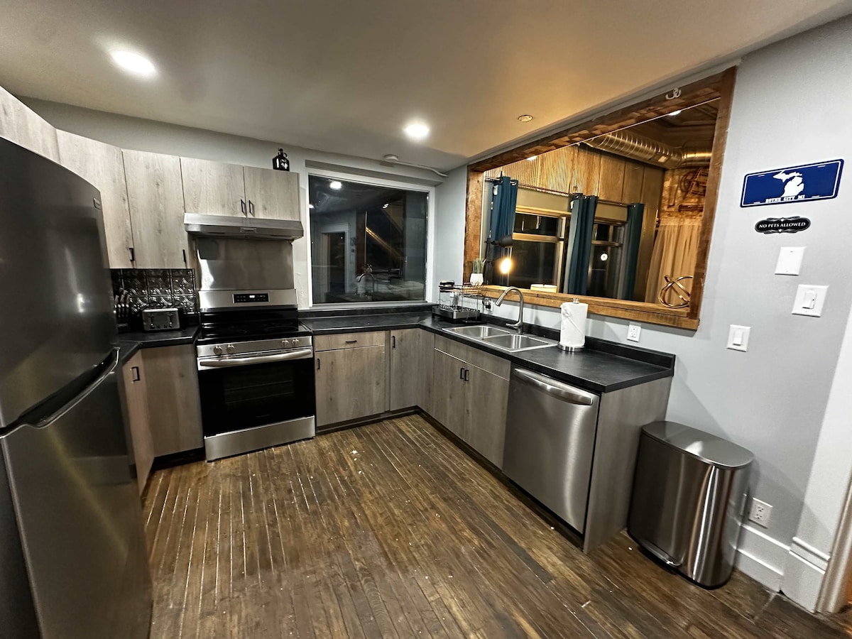 The kitchen features modern stainless steel appliances, including a refrigerator, stove, and dishwasher. Natural wood cabinetry contrasts with a dark countertop. A large window provides natural light, while the open layout allows easy movement and access to cooking spaces.
