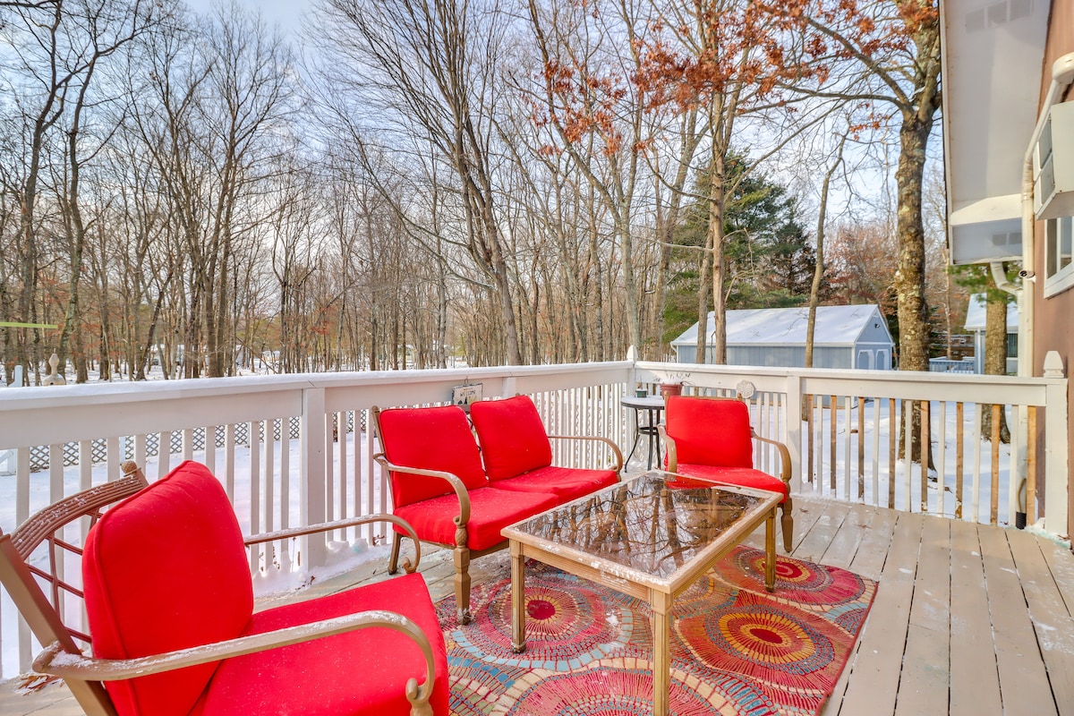 A deck area features a glass-top table surrounded by red cushioned chairs, inviting relaxation outdoors. The surrounding landscape showcases bare trees and a snowy yard, creating a serene setting. A small side table offers additional surface space.