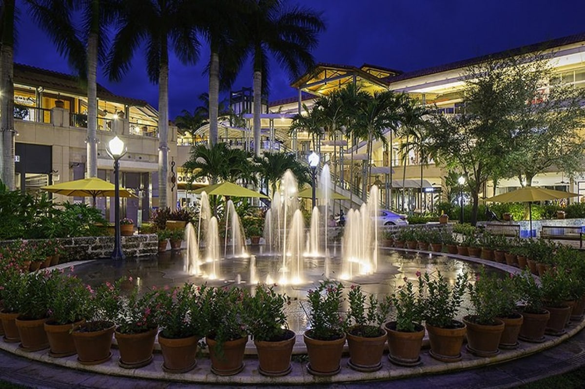 A tranquil courtyard is depicted with a circular fountain at its center, surrounded by potted plants and softly illuminated by lights. A lush palm tree backdrop adds to the serene ambiance, while warm lighting from the surrounding shops enhances the evening atmosphere.