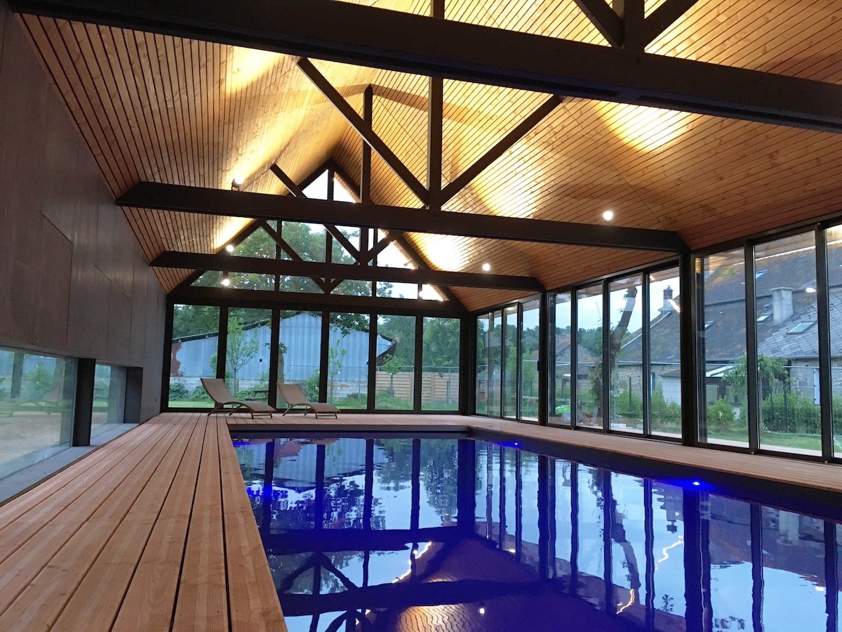 An indoor swimming pool is enclosed by large glass panels, allowing ample natural light to illuminate the space. Wooden beams extend across the ceiling, accentuating the modern design. Comfortable lounge chairs are positioned beside the water, providing a relaxing atmosphere.