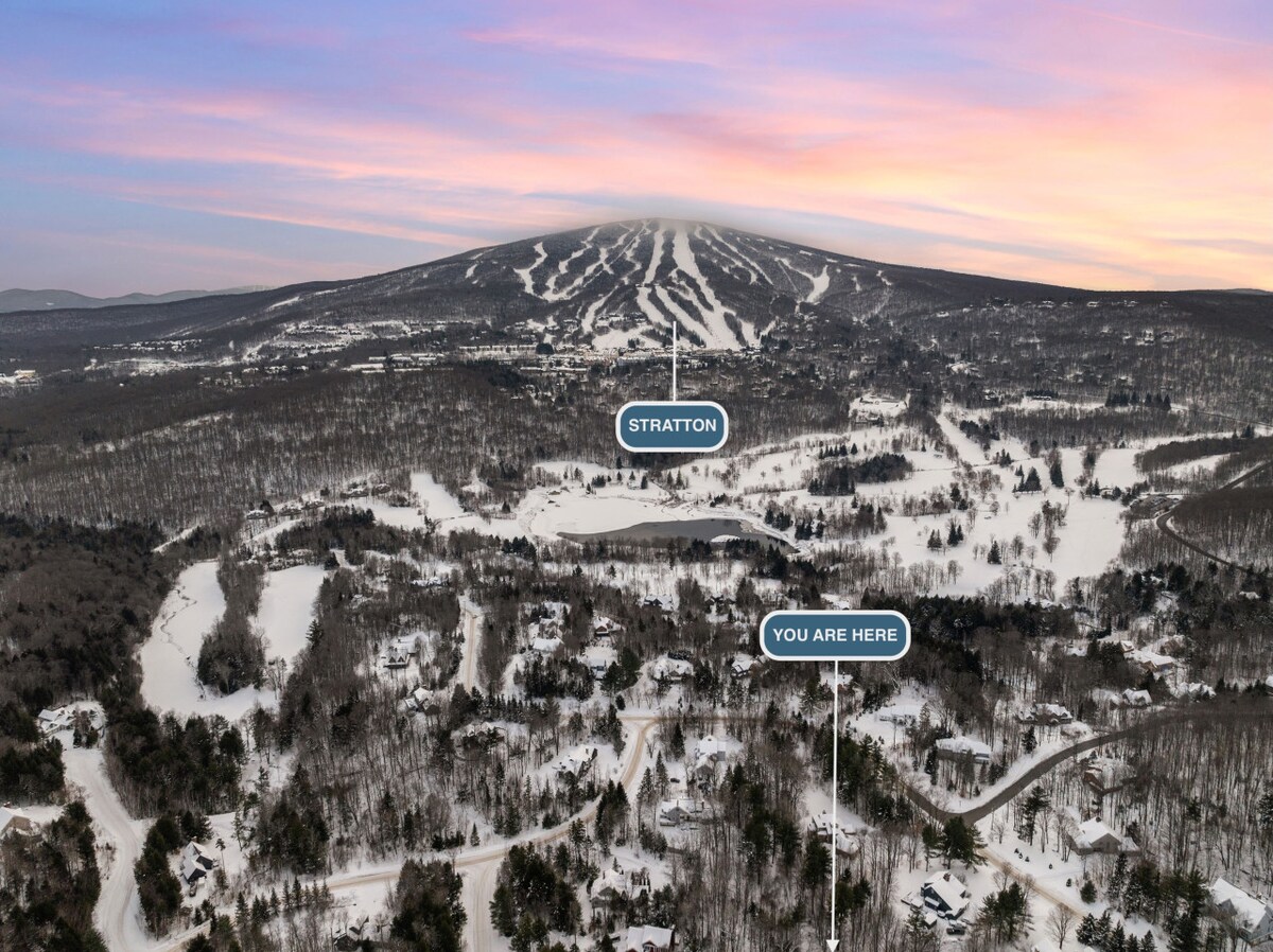 An aerial view showcases the snowy landscape surrounding Stratton Mountain. The slope of the mountain is visible in the background, with designated paths marked out. Tree-covered areas blend seamlessly with open spaces, providing a serene winter atmosphere.
